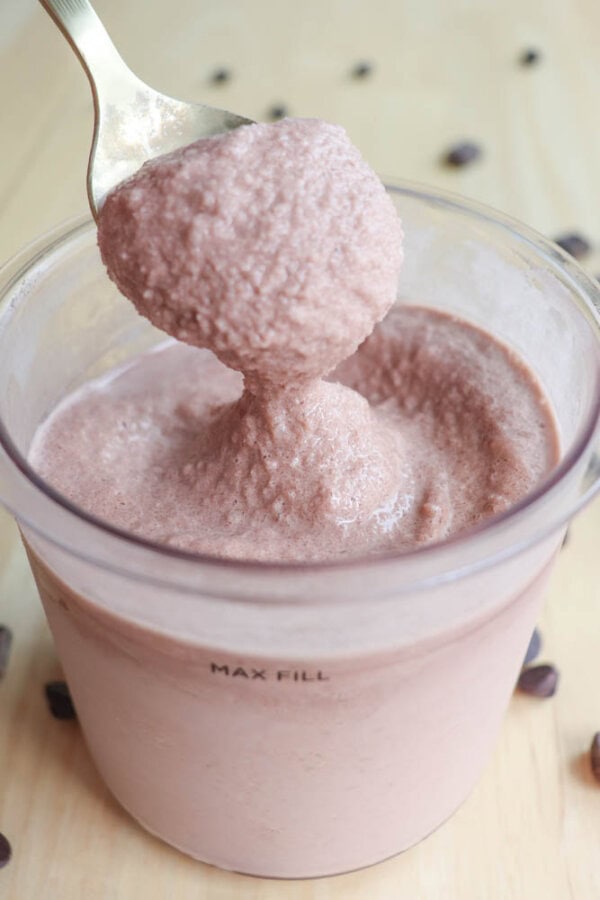 Spoon scooping thick strawberry smoothie from a transparent container. The smoothie appears creamy and textured, with a faint pinkish-brown color. The container has a MAX FILL mark and rests on a light wooden surface with scattered chocolate chips.