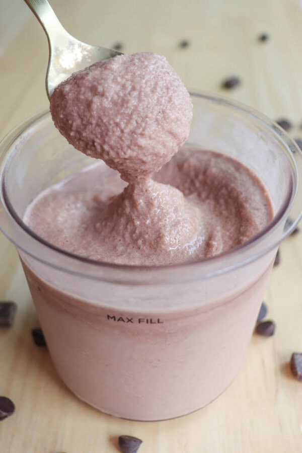 A spoon is lifting thick, frothy chocolate mousse from a transparent container marked MAX FILL. The mousse inside is creamy with a smooth texture. Scattered chocolate chips are visible on the wooden surface around the container.