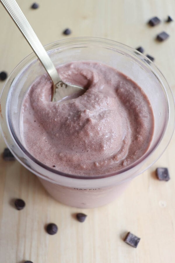 A cup filled with creamy chocolate ice cream, with a spoon inserted into it. Chocolate chips and chunks are scattered on the light wooden surface around the cup.