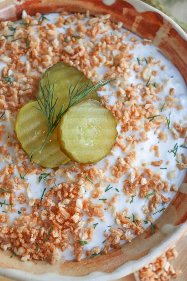 A bowl of ranch and dill dip topped with finely chopped fried onions, garnished with three pickle slices and a sprig of dill. The dip is served in a rustic ceramic bowl with a beige and brown pattern.
