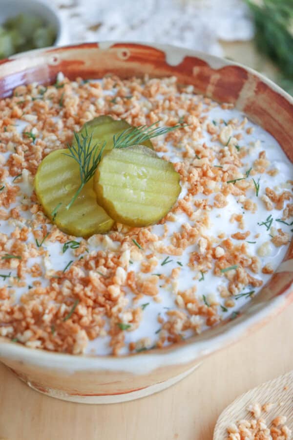 A bowl of salad topped with chopped nuts, garnished with three pickle slices and a sprig of dill. The creamy salad is presented in a rustic ceramic bowl on a wooden surface, with a spoon beside it.