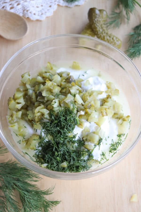 A clear glass bowl containing chopped pickles, fresh dill, and creamy white sauce on a wooden surface. Fresh dill sprigs and a pickle are scattered around, with a wooden spoon partially visible nearby.