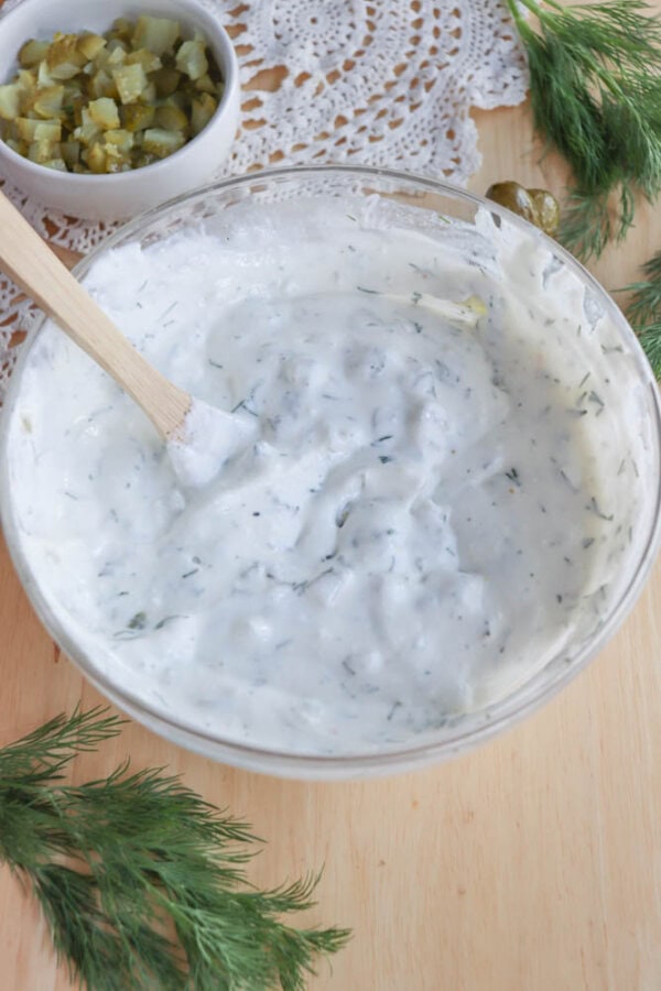 A bowl of creamy white sauce mixed with herbs, featuring a wooden spoon inside. Fresh dill is scattered around the bowl, and a small dish of chopped pickles is in the background, all placed on a light wooden surface with lace decor.
