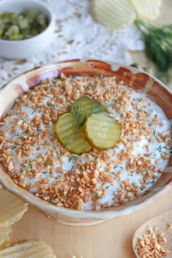 A bowl of creamy dip topped with crumbled crispy bits and garnished with sliced pickles and dill. A small bowl of pickles and potato chips are in the background on a wooden surface.