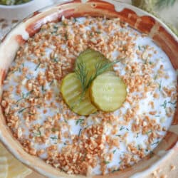 A bowl of creamy dip topped with fried onions, three pickle slices, and a sprig of dill. Crinkle-cut potato chips are on the side. A small bowl of chopped pickles is in the background.