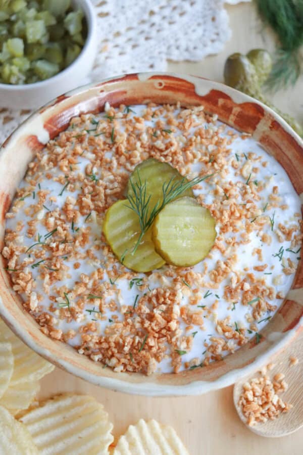 A bowl of creamy dip topped with fried onions, three pickle slices, and a sprig of dill. Crinkle-cut potato chips are on the side. A small bowl of chopped pickles is in the background.