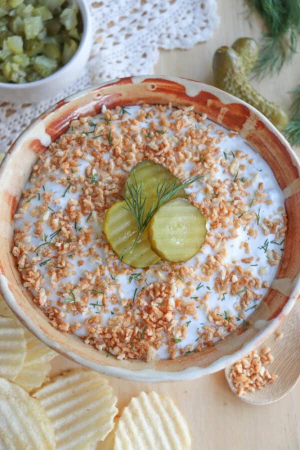 A bowl of dip topped with crispy fried onions, sliced pickles, and dill. The bowl is surrounded by ridged potato chips. A small dish of chopped pickles is in the background on a lace doily.