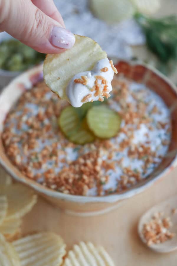 A hand holds a ridged potato chip dipped in a creamy white dip topped with crispy fried onions. The bowl in the background also contains the dip, garnished with pickles and more fried onions, surrounded by additional chips and a spoon.