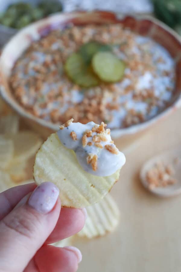 Close-up of a hand holding a ridged potato chip dipped in a creamy white sauce with crumbled toppings. A bowl in the background contains more dip, topped with sliced pickles and crumbles. Additional chips and a wooden spoon are nearby.