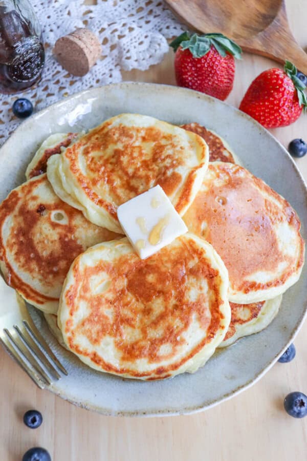 A plate of golden cottage cheese pancakes topped with a melting pat of butter. The wooden table is adorned with blueberries, strawberries, and a lace doily in the background. A fork is placed beside the plate.