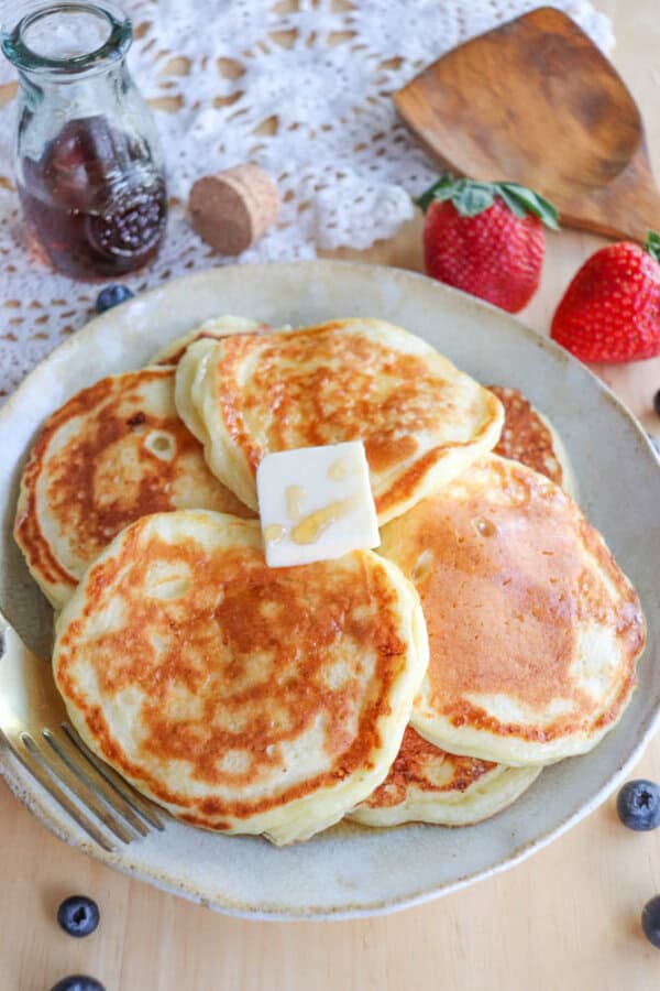 A plate of golden-brown cottage cheese pancakes topped with a pat of butter. Surrounding the plate are fresh strawberries, blueberries, a jar of syrup, and a fork. A wooden spatula and a lace tablecloth enhance this delightful scene.