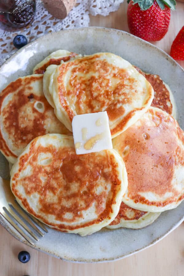 A plate of golden-brown cottage cheese pancakes topped with a pat of melting butter and a drizzle of honey. The plate is surrounded by fresh strawberries and blueberries, with a fork placed at the side.