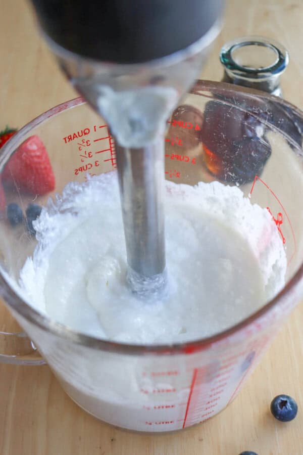 A hand blender is immersed in a glass measuring cup filled with a white creamy mixture, perfect for a cottage cheese pancake recipe. The cup is marked with red measurement lines. Fresh berries, including strawberries and blueberries, are scattered on the wooden surface nearby.