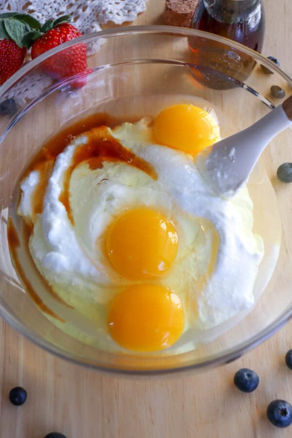 A glass bowl contains three egg yolks, sugar, yogurt, and vanilla extract being mixed with a spatula for a delightful cottage cheese pancake recipe. A strawberry, blueberries, and a small bottle of syrup are on the wooden surface nearby.