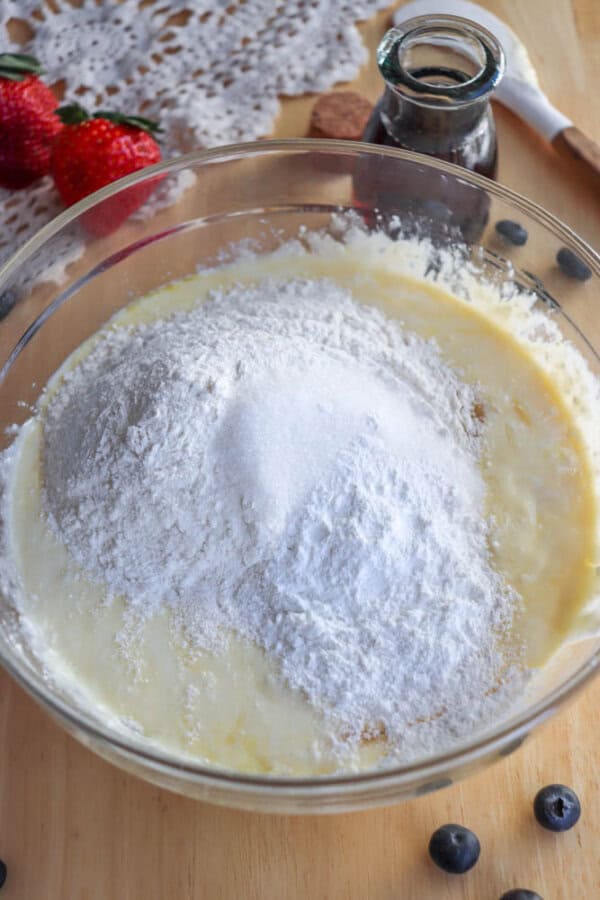 A glass bowl brimming with batter and a pile of flour hints at a delicious cottage cheese pancake recipe. Nearby are strawberries, a few blueberries, vanilla extract, a spoon, and a crocheted cloth on the wooden surface.