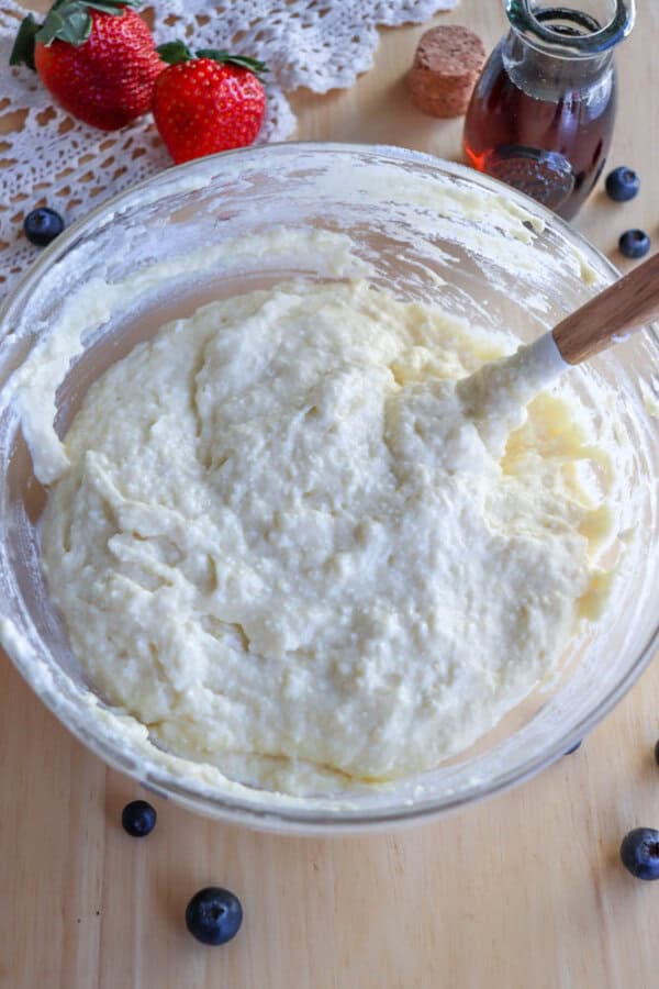 A bowl of batter for a delightful cottage cheese pancake recipe sits with a wooden spoon on a rustic wooden table. Fresh strawberries, blueberries, and a small glass bottle with a cork stopper add charm, while lace fabric completes the inviting scene.