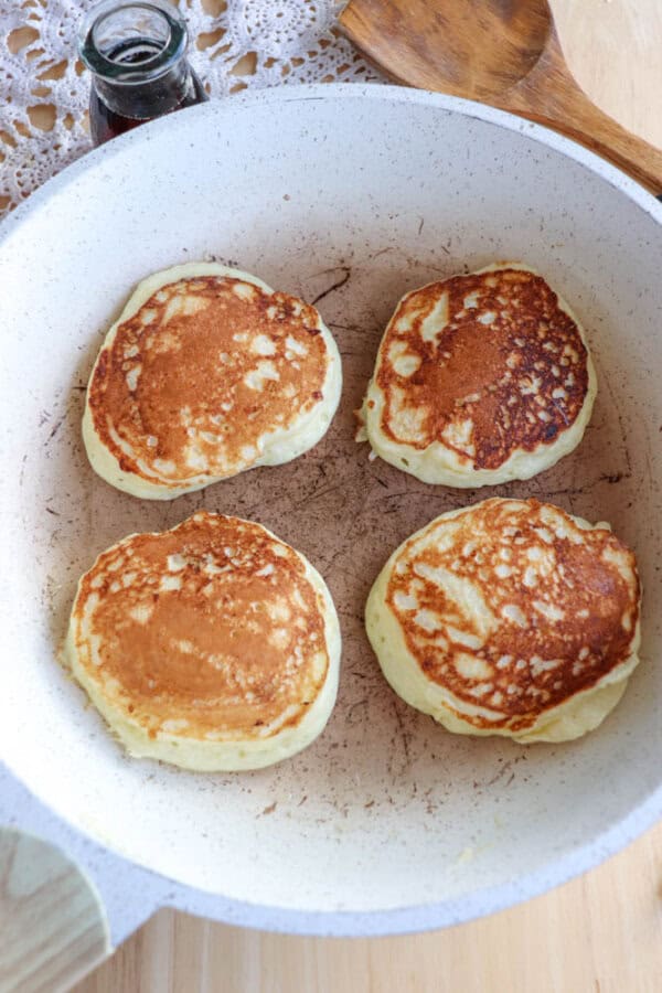 Four golden-brown cottage cheese pancakes sizzle in a white frying pan on a wooden surface. A wooden spatula and a small jar of syrup await nearby, with a decorative doily partially visible in the background, adding charm to this cozy scene.