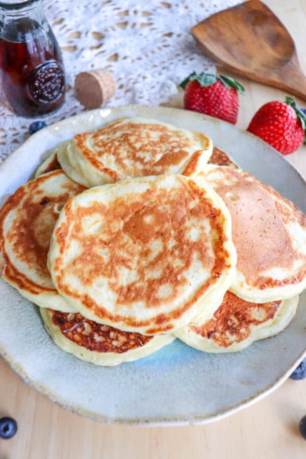 A plate of golden-brown cottage cheese pancakes stacked on a table with a doily. Next to the plate are fresh strawberries and a small bottle of syrup with a cork stopper. A wooden spatula is also visible in the background, ready to serve this delightful recipe.