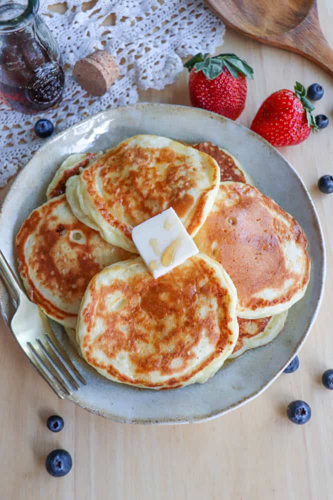 A plate of golden, fluffy pancakes made from a cottage cheese pancake recipe is topped with a pat of butter and drizzled with syrup. Fresh strawberries and blueberries are scattered around the plate, with a fork resting beside the pancakes on a wooden table.