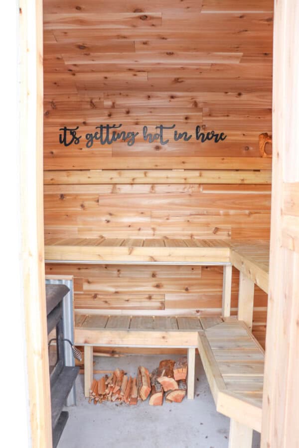 A small wooden sauna with benches and a firewood stack on the floor. A black wood stove is in the corner, and a sign reading its getting hot in here is mounted on the wood-paneled wall.