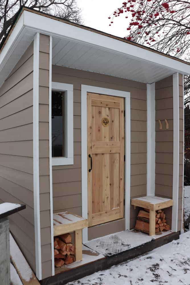 Small sauna with a wooden door, a small window to the left, and two log benches on either side. Firewood is stacked beneath the benches. The ground is lightly covered in snow, and bare tree branches are visible above.