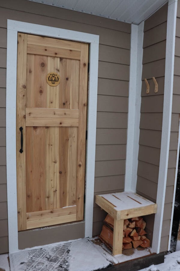 A wooden door with a round decoration is set in a brown wall with white trim. Next to the door is a small wooden stand holding cut firewood. Snow is on the ground, and two hooks are mounted on the wall above the stand.