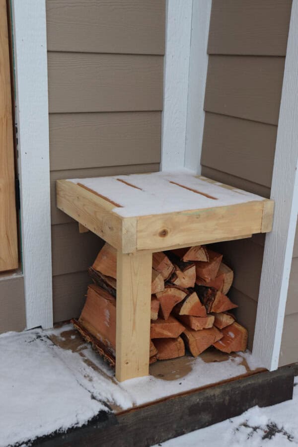 A stack of firewood neatly arranged under a simple wooden shelf attached to the corner of a house exterior. The wood is covered with a light layer of snow, and the ground around is dusted with snow.