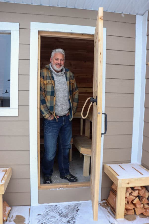 A man with gray hair and a beard, wearing a plaid jacket, gray sweater, and jeans, stands in the doorway of a small wooden building. Snow is on the ground, and firewood is stacked beside the entrance.