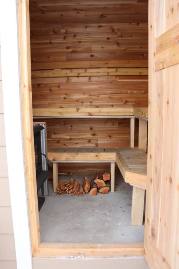 Wooden sauna interior with a bench along the right wall and wooden panels. Firewood is stacked on the floor beneath a small table. A heater is visible in the corner, and the door is partially open.