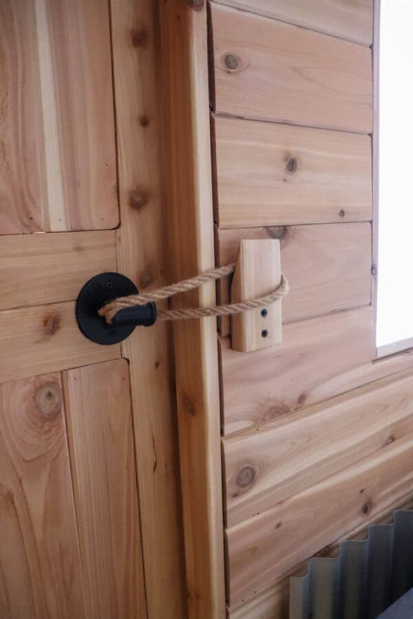 A wooden door with a rustic latch system. The door has a black metal handle, and a rope is used as a makeshift lock, tied between the handle and a wooden block on the frame. The wall and door have a natural wood finish.
