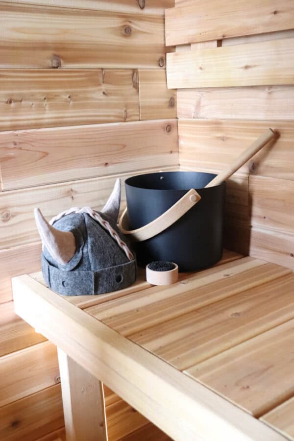 Wooden sauna interior with a felt hat featuring horns on a bench. A black sauna bucket with a ladle rests beside a small container. The setting suggests a Scandinavian sauna with traditional themes.