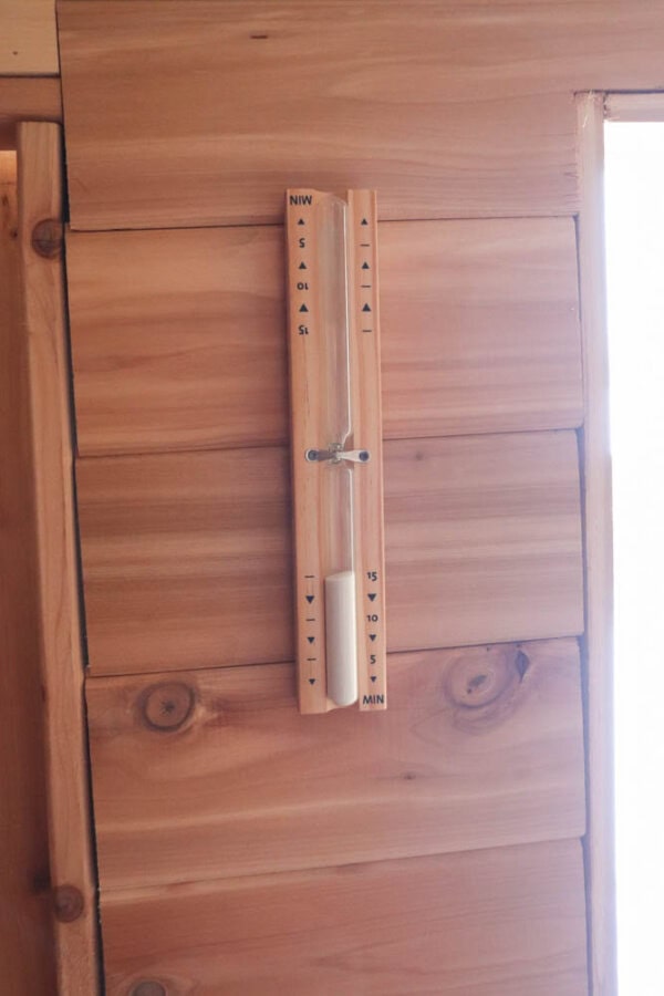 A wooden wall-mounted homopolar clock with a vertical rod and a sliding metal pointer. Time markers are inscribed on both sides of the rod, surrounded by light-colored wood paneling.