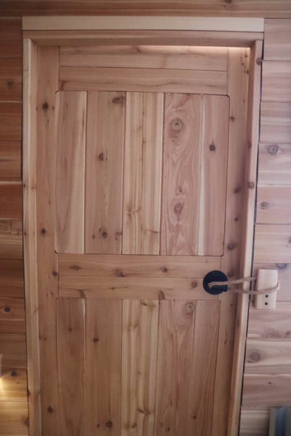 A wooden door with prominent grain patterns and a black metal handle. The door is set within a wooden frame, matching the paneled wall surrounding it. Soft lighting highlights the natural wood texture.