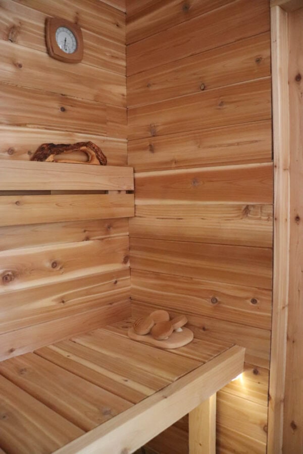 A cozy wooden sauna interior with a small corner bench. A clock hangs on the wall above, and a pair of wooden sandals rests on the bench. Soft lighting highlights the warm tones of the wood.