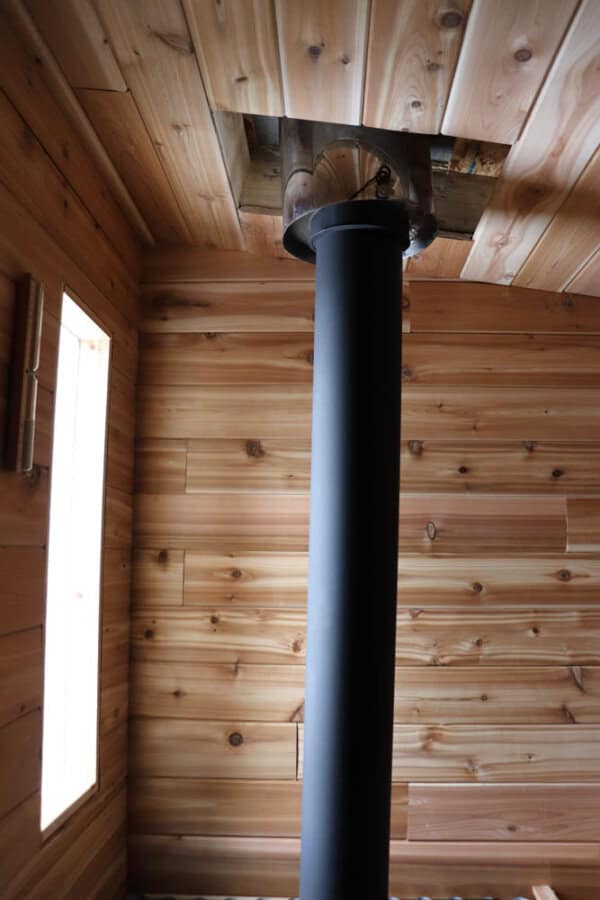 A black stove pipe extends vertically through a wooden ceiling in a room with wooden walls. A rectangular window is visible on the left, allowing natural light to illuminate the warm wood tones.