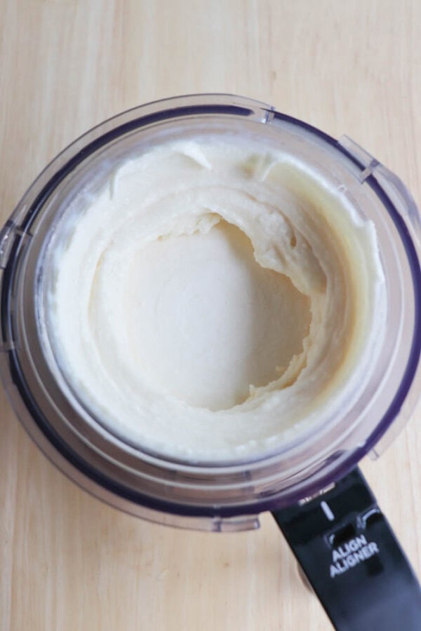 Top view of a food processor bowl containing smooth, creamy mashed potatoes. The mixture is light in color and the sides of the bowl show marks from blending. The countertop is light wood.