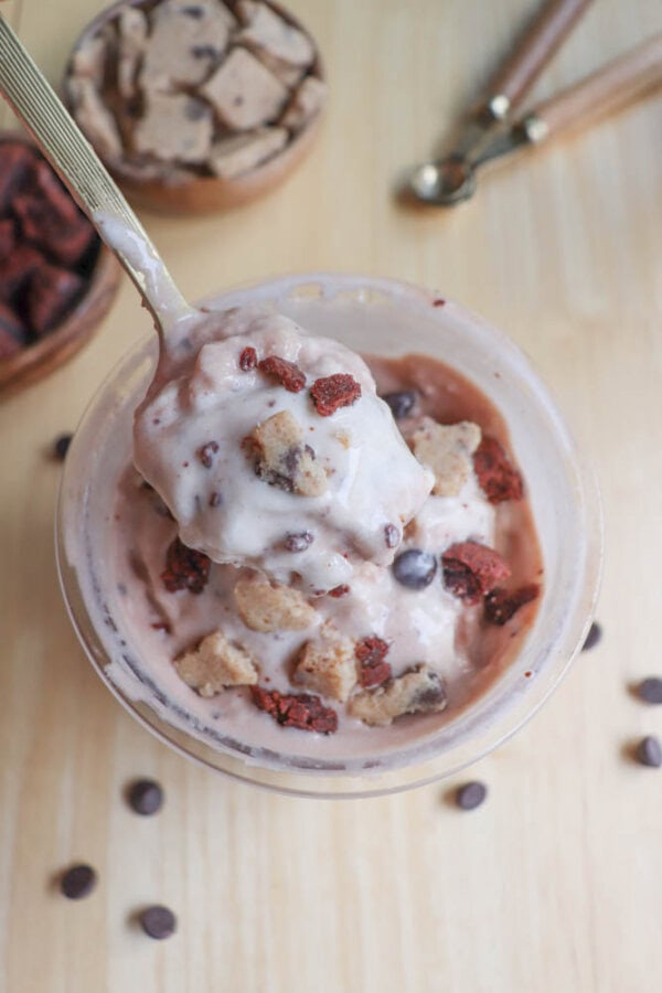 A cup of swirled ice cream topped with chunks of cookie dough, chocolate chips, and brownie pieces on a wooden table. A gold spoon is scooping up a portion. Two small bowls with more toppings are in the background.