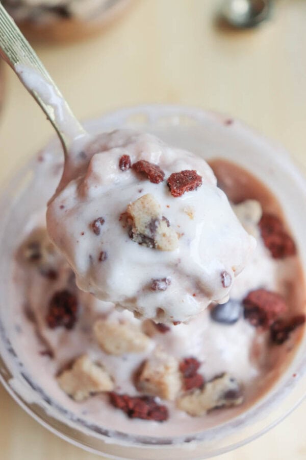 A spoonful of creamy ice cream, mixed with chocolate chunks and cookie dough pieces, is lifted above a container filled with more ice cream and toppings. The background is slightly blurred, emphasizing the desserts texture and ingredients.