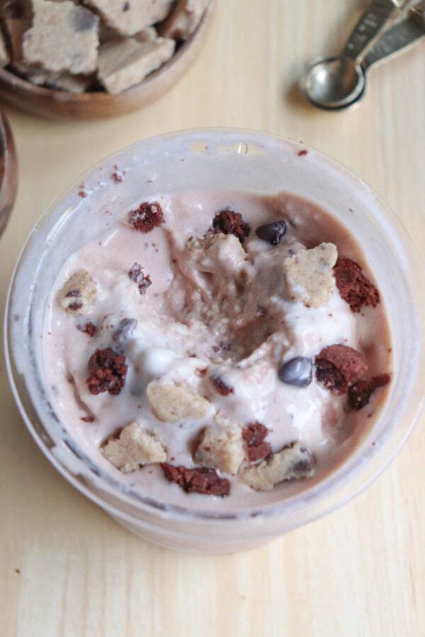 A bowl of creamy ice cream topped with chunks of chocolate chip cookie dough and chocolate pieces. The ice cream is swirled with chocolate and vanilla flavors, and measuring spoons are visible in the background on a wooden surface.