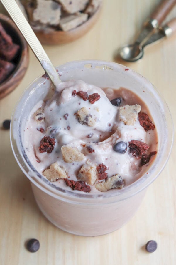 A container of ice cream topped with cookie chunks and chocolate bits, with a spoon dipped in. In the background, there are bowls with more cookie pieces and an ice cream scoop on a wooden surface.