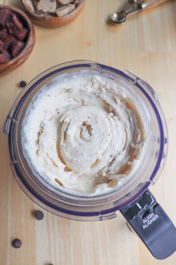 Top view of a blender filled with creamy banana ice cream on a wooden surface. Surrounding the blender are small bowls containing banana slices and cocoa powder. Chocolate chips are scattered nearby.