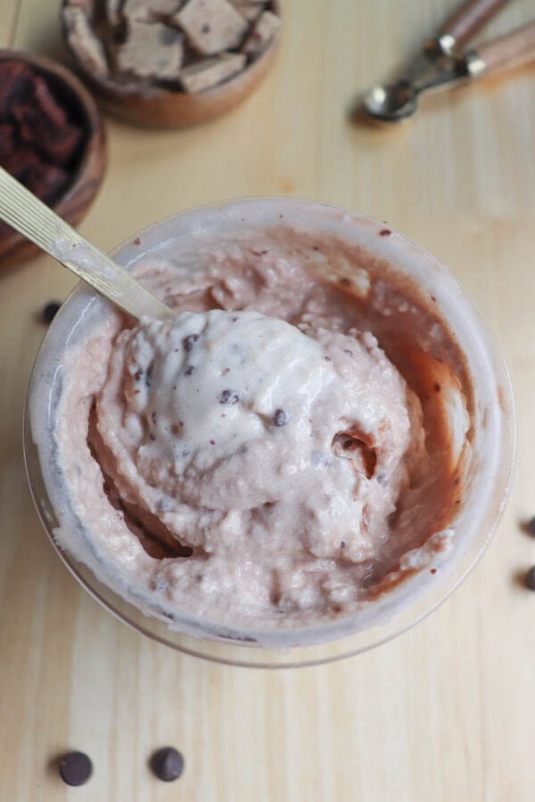 A bowl of creamy ice cream with chocolate chips, a spoon placed inside. The ice cream has a light brown color, and small chocolate chips are sprinkled around the bowl. Wooden texture surface and bowls with ingredients in the background.