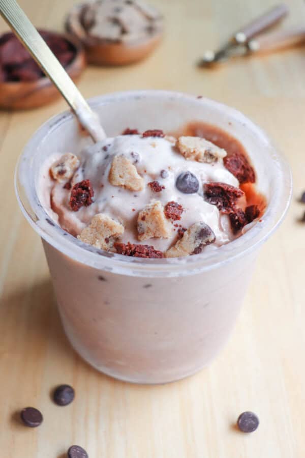 A cup of creamy ice cream topped with chocolate chips, cookie dough chunks, and brownie pieces, with a spoon sticking out. Scattered chocolate chips are on the wooden surface around the cup.