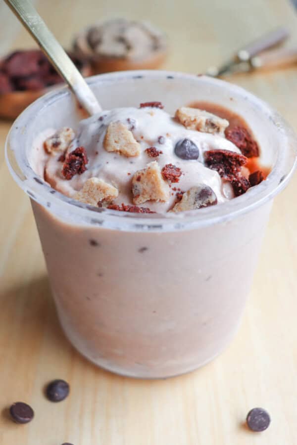 Cup of creamy dessert topped with cookie dough chunks and chocolate chips, served with a spoon. The smooth, thick texture and chocolate pieces are visible. The background is a light wooden surface with blurred utensils.