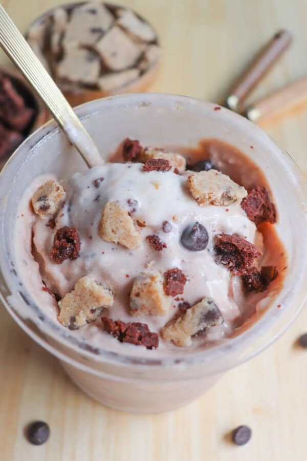 A cup filled with chocolate and vanilla swirl ice cream topped with chunks of cookie dough, brownies, and chocolate chips. A spoon is placed in the ice cream, and a lid with more cookie dough pieces is visible in the background.