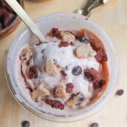 A bowl of ice cream topped with chunks of cookie dough and chocolate pieces. A spoon is sticking out of the bowl. In the background are small bowls with more cookie dough chunks and chocolate pieces on a wooden surface.