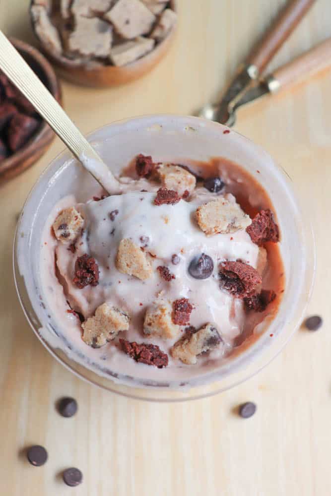 A bowl of ice cream topped with chunks of cookie dough and chocolate pieces. A spoon is sticking out of the bowl. In the background are small bowls with more cookie dough chunks and chocolate pieces on a wooden surface.