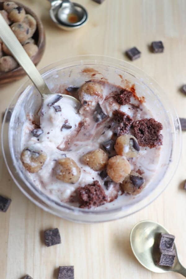 Bowl of chocolate and vanilla ice cream topped with cookie dough balls, brownie chunks, and chocolate pieces. A spoon rests in the bowl, with scattered chocolate chunks around it on a wooden surface.