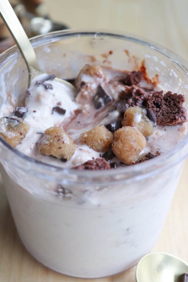 A cup filled with creamy dessert, topped with chunks of chocolate brownie, chocolate chips, and small cookie dough bites. A spoon is dipped into the dessert, resting on the edge of the cup. The background is a light wooden surface.
