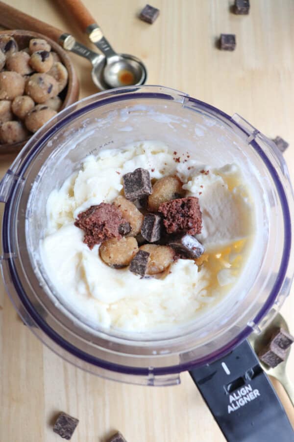 A top-down view of a blender filled with ingredients for ice cream. The mixture includes vanilla ice cream, cookie dough chunks, and chocolate pieces. A small bowl with more cookie dough and a measuring spoon are nearby on a wooden surface.